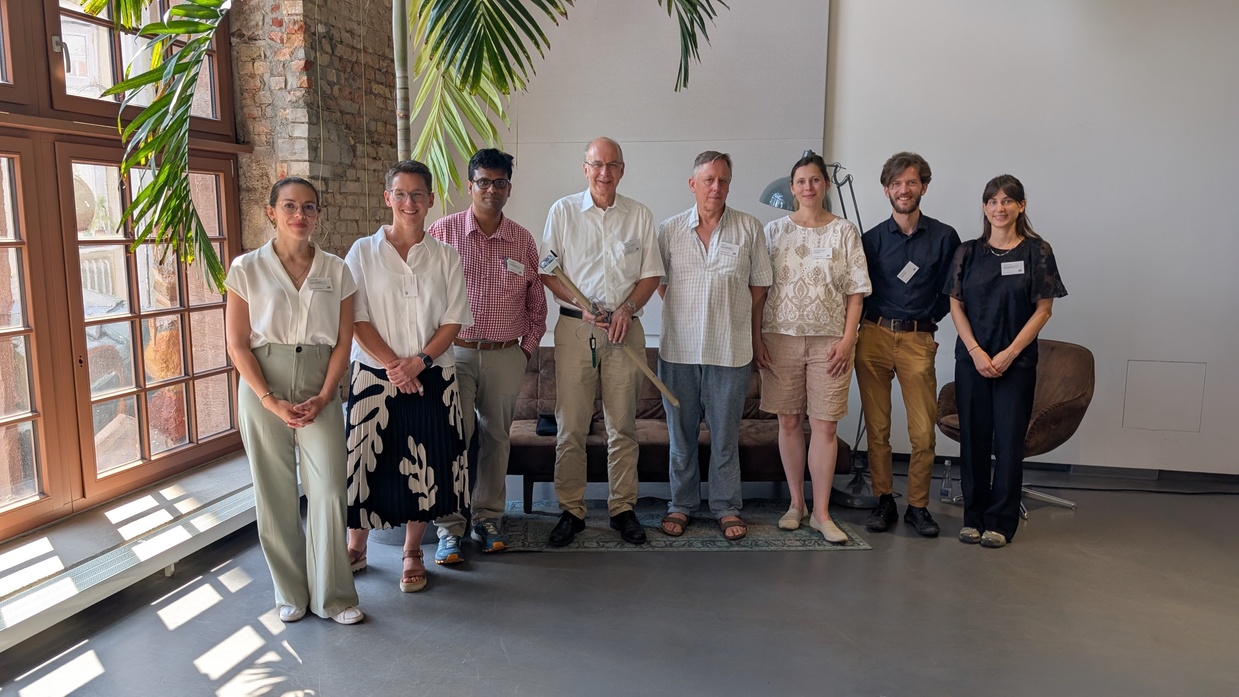 Foto von acht Personen, die nebeneinander in einer Reihe stehen. Die vierte Person von links hält eine Bodensonde in der Hand. Am oberen Bildrand hängen Palmenblätter in das Foto. Hinter der Gruppe steht ein braunes Sofa.